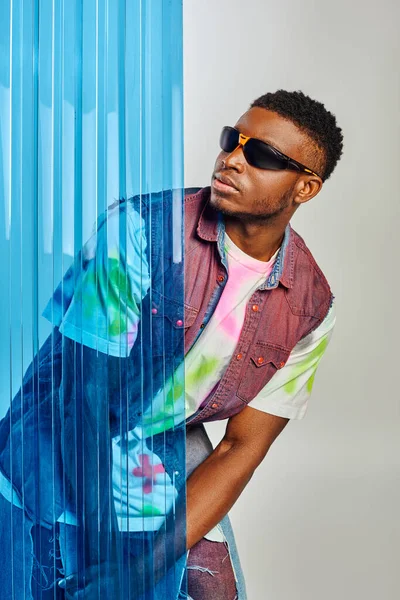 Modelo afroamericano confiante em óculos de sol, colete jeans jeans jeans e rasgado em pé atrás de folha de policarbonato azul em fundo cinza, tiro de moda, roupas DIY, estilo de vida sustentável — Fotografia de Stock