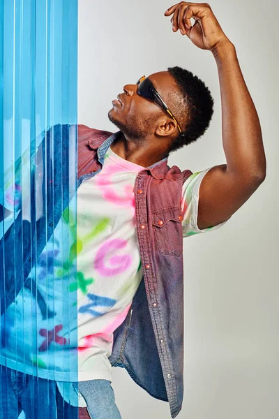 Seitenansicht des trendigen jungen Afroamerikaners mit Sonnenbrille, bunter Jeansweste und T-Shirt posiert und steht hinter blauem Polycarbonat-Laken auf grauem Hintergrund, DIY-Kleidung, nachhaltiger Lebensstil — Stockfoto