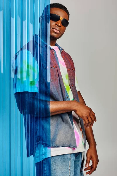 Jeune modèle afro-américain masculin en lunettes de soleil, gilet en denim et t-shirt coloré posant et debout derrière une feuille de polycarbonate bleu sur fond gris, vêtements de bricolage, mode de vie durable — Photo de stock