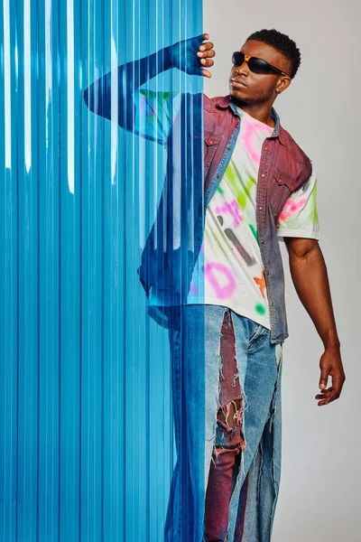 Bonito modelo afroamericano joven en gafas de sol, chaleco de mezclilla y camiseta tocando la hoja de policarbonato azul y de pie sobre fondo gris, sesión de moda, ropa de bricolaje, estilo de vida sostenible — Stock Photo