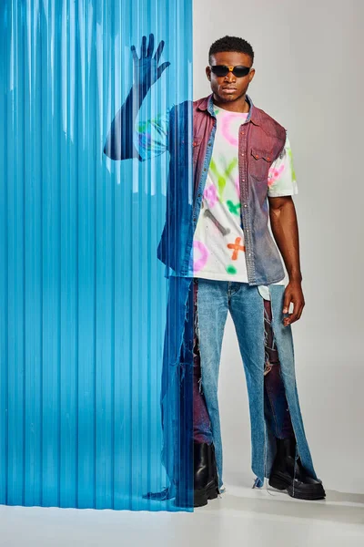 Modèle afro-américain masculin en gilet en denim pleine longueur, jean déchiré et lunettes de soleil touchant une feuille de polycarbonate bleu et debout sur fond gris, vêtements DIY, mode de vie durable — Photo de stock