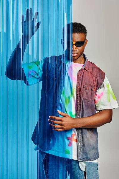Modèle afro-américain masculin tendance en lunettes de soleil, t-shirt coloré et gilet en denim posant derrière une feuille de polycarbonate bleu sur fond gris, tournage de mode, vêtements DIY, mode de vie durable — Photo de stock