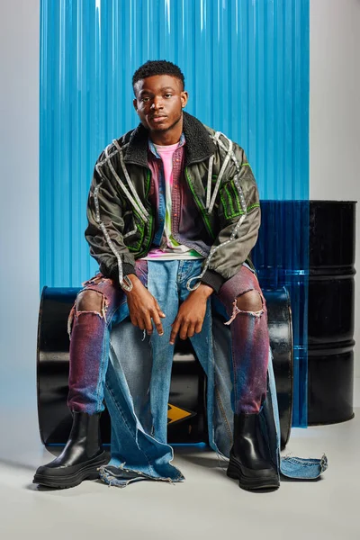 Trendy young afroamerican man in outwear jacket with led stripes and ripped jeans sitting on fuel barrel and looking at camera near blue polycarbonate sheet on grey background, DIY clothing — Stock Photo