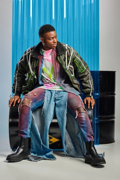 Stilvoller afroamerikanischer Mann in Outwear-Jacke mit Bleistreifen und bunt zerrissenen Jeans sitzt auf einem Benzinfass neben blauem Polycarbonat-Laken und auf grauem Hintergrund, DIY-Kleidung, nachhaltiger Lebensstil — Stockfoto