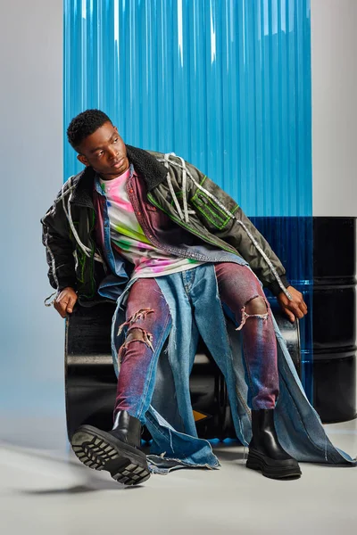 Jeune afro-américain confiant modèle masculin en jeans déchiré et veste de vêtements avec des rayures led regardant loin tout en étant assis sur le baril de carburant près de la feuille de polycarbonate bleu sur gris, mode de vie durable — Photo de stock