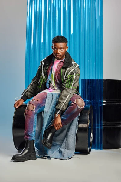 Впевнена молода афроамериканська модель з модною зачіскою в куртці для взуття зі свинцевими смугами та рваними джинсами, сидячи на паливній бочці поблизу синього полікарбонату на сірому, одяг DIY — стокове фото