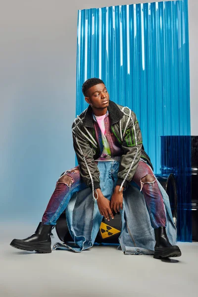 Stylish young afroamerican man in modern ripped jeans and outwear jacket with led stripes sitting on fuel barrel near blue polycarbonate sheet on grey background, DIY clothing, sustainable lifestyle — Stock Photo
