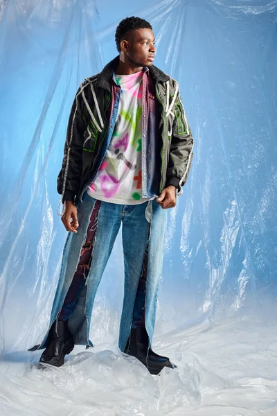 Pleine longueur de confiant jeune homme afro-américain en veste de vêtements avec des rayures led et jeans déchiré regardant loin tout en se tenant près de cellophane brillant sur fond bleu, mode de vie durable — Photo de stock