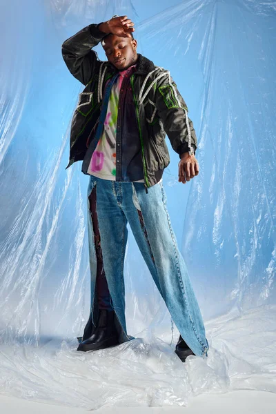 Modelo afroamericano joven y atractivo en chaqueta de outwear con rayas led y jeans rasgados tocando la frente y de pie sobre celofán brillante sobre fondo azul, atuendo urbano y ropa de bricolaje - foto de stock