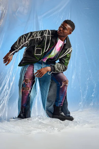 Modelo afroamericano jovem elegante em jaqueta de roupas com listras led e jeans rasgados olhando para longe e posando em celofane brilhante em fundo azul, roupa urbana e estilo de vida sustentável — Fotografia de Stock
