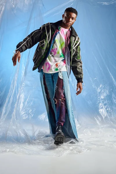 Homem afroamericano jovem na moda em jaqueta de roupas com listras led e jeans rasgados andando e olhando para a câmera em celofane brilhante no fundo azul, roupa urbana, roupas DIY — Fotografia de Stock
