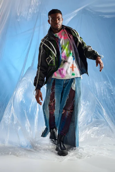 Pleine longueur de confiance jeune modèle afroaméricain en veste de vêtements de dessus avec des rayures led et jeans déchiré marchant sur cellophane brillant sur fond bleu, tenue urbaine, vêtements de bricolage — Photo de stock