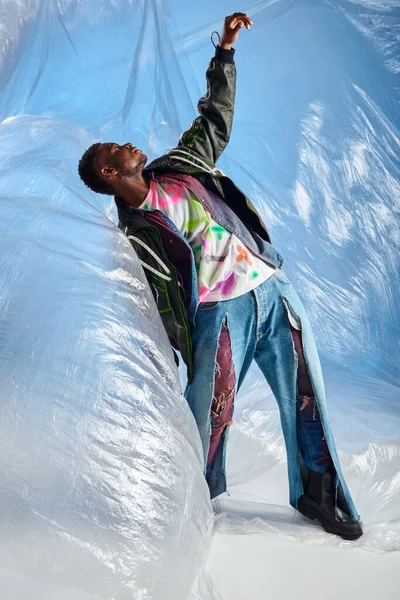 Вид збоку добре виглядає на афроамериканську чоловічу модель в піджаку з світлодіодними смугами та рваними джинсами, що стоять на глянцевому мольберті на синьому фоні, одязі DIY, стійкому способі життя — стокове фото
