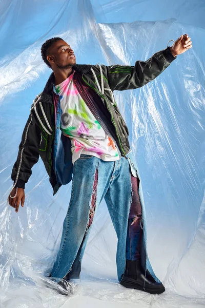 Модний афроамериканський чоловік у куртці з світлодіодними смугами та рваними джинсами позує на глянцевому целофані на синьому фоні, міському вбранні та сучасній позі, одязі, стійкому способі життя — стокове фото
