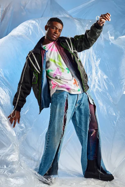 Pleine longueur d'homme afro-américain confiant en veste de sport avec des rayures led et un jean déchiré regardant la caméra près de cellophane brillant sur fond bleu, pose moderne, mode de vie durable — Photo de stock