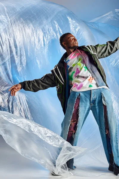 Trendy junges afroamerikanisches Model posiert in Outdoor-Jacke mit Bleistreifen und zerrissenen Jeans auf glänzendem Zellophan auf blauem Hintergrund, urbanem Outfit, DIY-Kleidung, nachhaltigem Lebensstil — Stockfoto