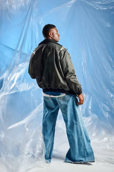 Vista lateral de hombre afroamericano confiado y elegante en chaqueta de outwear con rayas led y jeans de pie cerca de celofán sobre fondo azul, traje urbano y expresión creativa, ropa de bricolaje - foto de stock