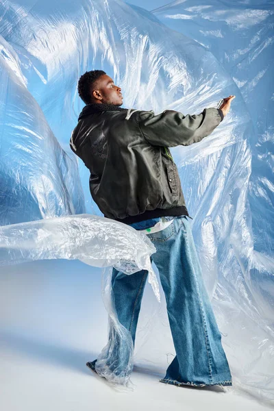 Vue latérale du jeune modèle afro-américain masculin à la mode en veste de sport avec rayures et jeans à led posant sur un cellophane brillant sur fond bleu, tenue urbaine, expression créative, vêtements de bricolage — Photo de stock