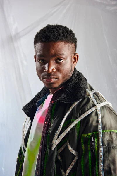 Portrait of young afroamerican model in outwear jacket with led stripes looking at camera near glossy cellophane on grey background, urban outfit and modern pose, creative expression, DIY clothing — Stock Photo