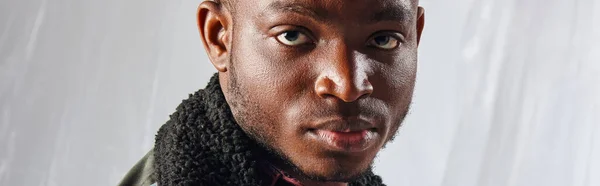 Portrait of confident young afroamerican man in outwear jacket looking at camera while standing near glossy cellophane on grey background, urban outfit and modern pose, banner, creative expression — Stock Photo