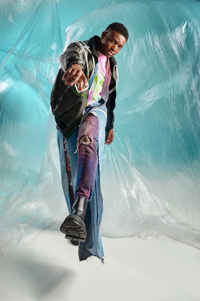 Voller Länge trendiges junges afroamerikanisches Model in Outwear-Jacke und zerrissenen Jeans, das in die Kamera schaut und in der Nähe von glänzendem Zellophan auf türkisfarbenem Hintergrund spaziert, kreativer Ausdruck, DIY-Kleidung — Stockfoto