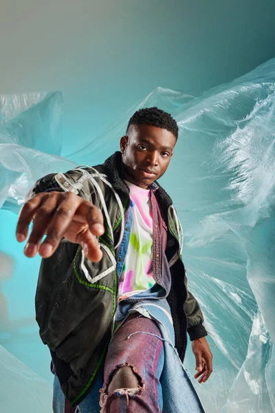 Portrait of confident young afroamerican man in outwear jacket and ripped jeans posing with cellophane on turquoise background, urban outfit and modern pose, creative expression, DIY clothing — Stock Photo