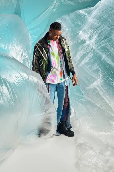 Повна довжина модної афроамериканської чоловічої моделі в одязі піджака зі світлодіодними смугами та рваними джинсами, що стоять біля целофана на бірюзовому фоні, творчий вираз, одяг DIY — стокове фото