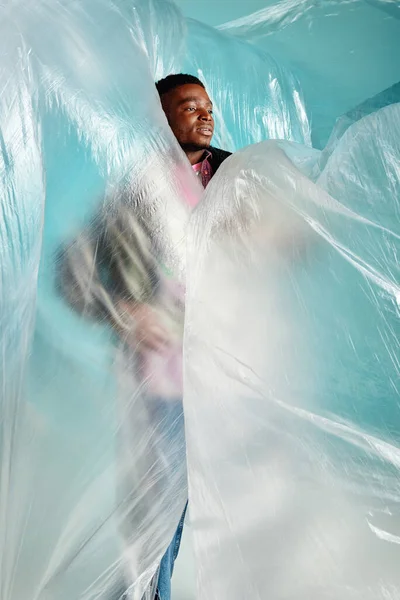 Junger afrikanisch-amerikanischer Mann mit trendiger Frisur, der wegschaut, während er in Zellophan auf türkisfarbenem Hintergrund steht, urbanes Outfit und moderne Pose, kreativer Ausdruck, DIY-Kleidung — Stockfoto