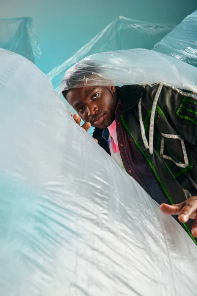 Porträt eines selbstbewussten jungen afroamerikanischen männlichen Modells in Unterhosenjacke mit LED-Streifen, das in die Kamera schaut, in der Nähe von glänzendem Zellophan auf türkisfarbenem Hintergrund, kreativer Ausdruck, DIY-Kleidung — Stockfoto