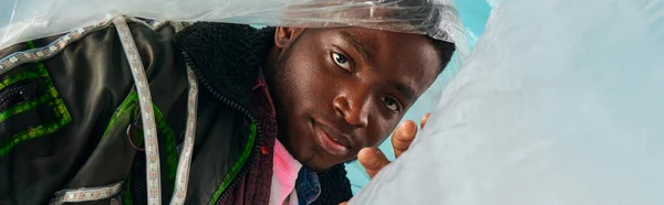 Portrait de jeune homme afro-américain élégant en veste de vêtements avec des rayures led posant avec cellophane brillant sur fond turquoise, tenue urbaine et pose moderne, bannière, expression créative — Photo de stock