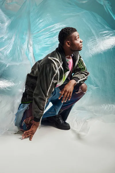 Vista lateral do homem afroamericano bem parecido em jaqueta de roupas com listras led e jeans rasgados olhando para cima perto de celofane brilhante em fundo turquesa, expressão criativa, roupas DIY — Fotografia de Stock