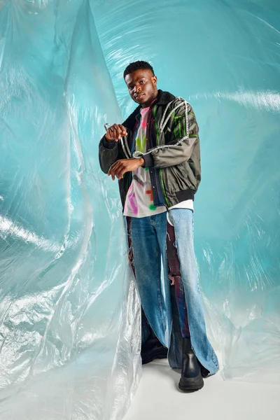 Comprimento total do homem afroamericano elegante em jeans rasgados tocando manga de jaqueta de roupas com listras led perto de celofane em fundo turquesa, roupa urbana e pose moderna, roupas DIY — Stock Photo