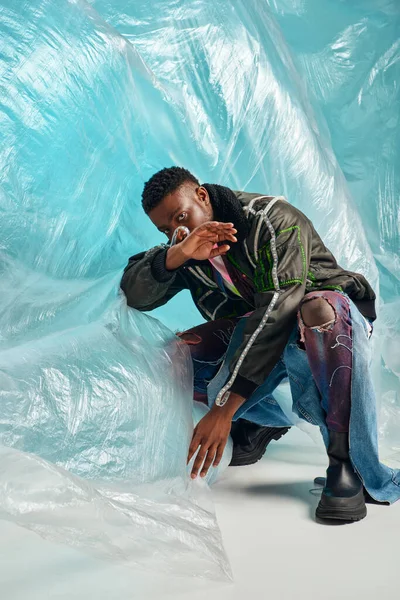 Expresión creativa, ropa de bricolaje, modelo joven afroamericano en chaqueta de outwear con rayas led y vaqueros de moda posando y cubriendo la cara cerca de celofán sobre fondo turquesa - foto de stock