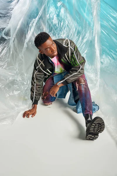Selbstbewusstes afrikanisch-amerikanisches Model in Outwear-Jacke mit Bleistreifen und zerrissenen Jeans, die in der Nähe von glänzendem Zellophan auf türkisfarbenem Hintergrund wegschaut, urbanem Outfit und moderner Pose, DIY-Kleidung — Stockfoto