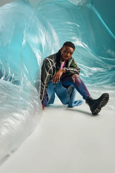 Jovem modelo afroamericano em jaqueta de roupas com listras led e jeans rasgados sentados perto de celofane brilhante em fundo turquesa, roupa urbana e pose moderna, expressão criativa, roupas DIY — Fotografia de Stock
