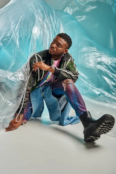 Modelo masculino americano africano na moda em jeans rasgados e jaqueta de roupas com listras led olhando para longe e posando perto de celofane em fundo turquesa, expressão criativa, roupas DIY — Fotografia de Stock