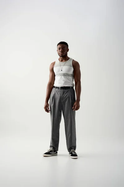 Junges und selbstbewusstes afroamerikanisches Model in ärmellosem T-Shirt und Hose, das auf grauem Hintergrund in die Kamera blickt, zeitgenössisches Shooting mit stilvoller Kleidung, lässiger Kleidung — Stockfoto