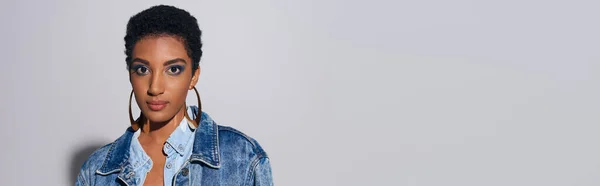 Retrato de moda jovem modelo afro-americano com maquiagem ousada em jaqueta jeans e brincos dourados olhando para a câmera em fundo cinza com espaço de cópia, conceito de moda jeans, banner — Fotografia de Stock