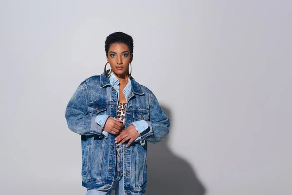 Модная африканская американка с короткими волосами, смотрящая в камеру, позируя в джинсовой куртке и золотых серьгах на сером фоне, джинсовая концепция моды — стоковое фото
