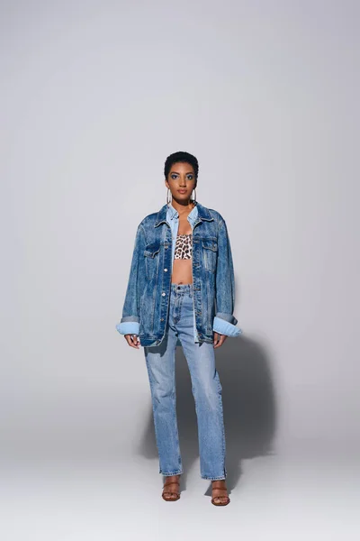 Полная длина модной африканской женщины с короткими волосами и смелым макияжем, позируя в джинсовой куртке и джинсах, стоя на сером фоне, джинсовой концепции моды — стоковое фото