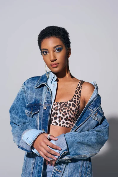 Портрет модной и уверенной в себе африканской женщины с смелым макияжем, позирующей сверху с изображением животных и джинсовой куртки на сером фоне, концепция джинсовой моды — стоковое фото