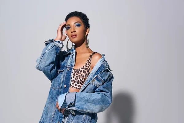 Confident young african american model with bold makeup touching short hair while posing in denim jacket and top with animal print on grey background, denim fashion concept — Stock Photo