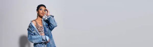 Jovem afro-americana de cabelos curtos com maquiagem ousada e brincos dourados tocando a cabeça enquanto posando em cima e jaqueta de ganga em fundo cinza, conceito de moda jeans, banner — Fotografia de Stock