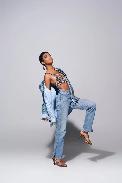 Полная длина модной африканской американкой с смелым макияжем позирует в верхней части с животным принтом, джинсовой курткой и джинсы, стоя на сером фоне, джинсовой концепции моды — стоковое фото