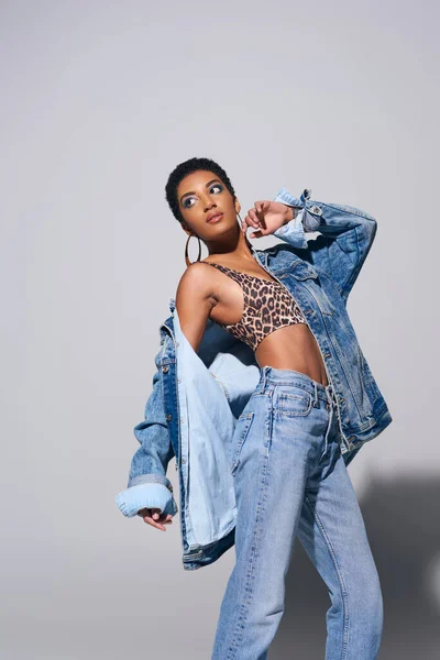 Модна афроамериканська модель з коротким волоссям, яскравим макіяжем і золотими сережками в джинсовій куртці, зверху і джинсах, стоячи на сірому фоні, концепція джинсової моди — стокове фото