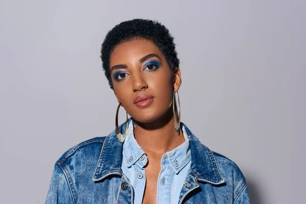 Portrait of modern and confident young african american woman with bold makeup and golden earrings wearing denim outfit and looking at camera on grey background, denim fashion concept — Stock Photo