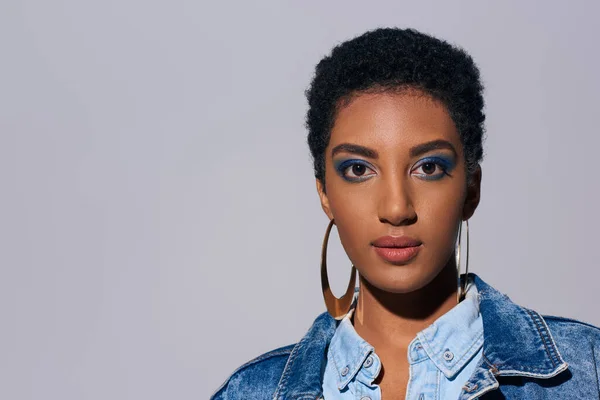Selbstbewusste junge afrikanisch-amerikanische Frau mit lebhaftem Make-up und goldenen Ohrringen im Jeans-Outfit, die isoliert auf graues Denim-Modekonzept in die Kamera blickt — Stockfoto