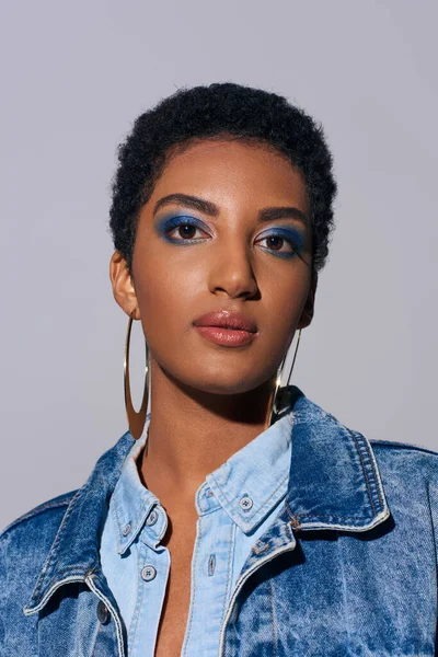 Portrait de femme afro-américaine branchée avec un maquillage audacieux et des boucles d'oreilles dorées posant en tenue denim et regardant la caméra isolée sur un concept de mode gris denim — Photo de stock