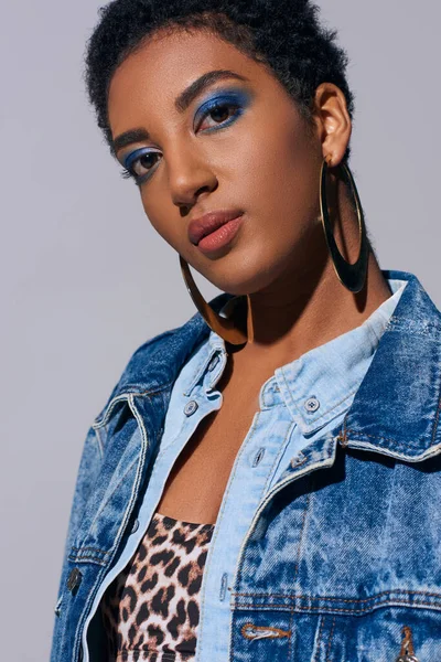 Confident and short haired african american woman with bold makeup and golden earrings wearing top and denim outfit while standing isolated on grey, denim fashion concept — Stock Photo