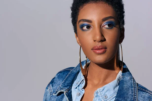 Portrait of stylish and short haired african american young woman with vivid makeup and denim jacket looking away while standing isolated on grey, denim fashion concept — Stock Photo